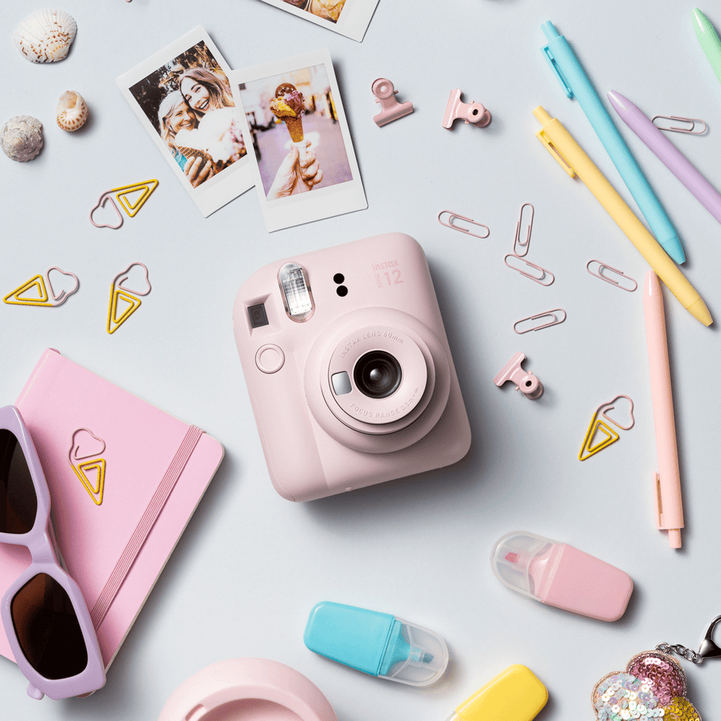 Sfeerafbeelding instax mini 12™ camera Blossom Pink. Op de foto ligt de camera op een pastel blauwe achtergrond met accessoires zoals: paperclips, pennen, highlighters, foto's, zonnebril en een notebook. 