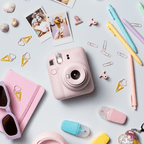 Sfeerafbeelding instax mini 12™ camera Blossom Pink. Op de foto ligt de camera op een pastel blauwe achtergrond met accessoires zoals: paperclips, pennen, highlighters, foto's, zonnebril en een notebook. 