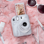 Sfeerafbeelding instax mini 12™ camera Clay White.  De camera ligt op een roze achtergrond met accessoires als: make-up kwasten, zonnebril, oorbellen en nagellak.Uit de camera komt een foto van een hand. 
