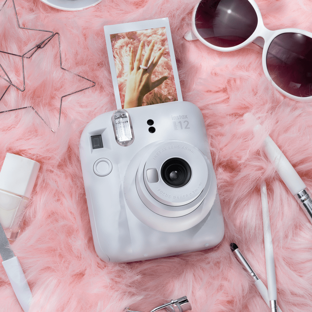Sfeerafbeelding instax mini 12™ camera Clay White.  De camera ligt op een roze achtergrond met accessoires als: make-up kwasten, zonnebril, oorbellen en nagellak.Uit de camera komt een foto van een hand. 