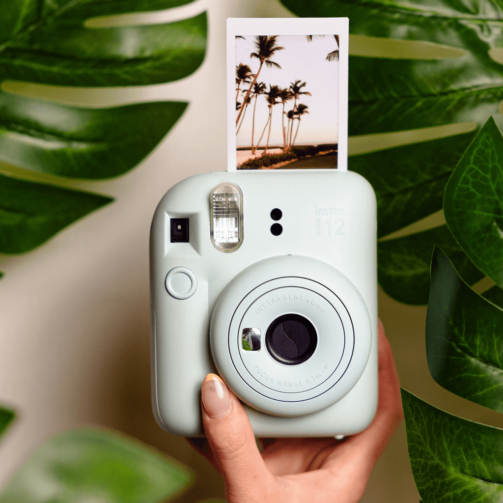 Sfeerafbeelding instax mini 12™ camera Mint Green, waarop de camera te zien is op de voorgrond en op de achtergrand bladeren van een plant zichtbaar zijn. Uit de camera komt een afbeelding met palmbomen. 