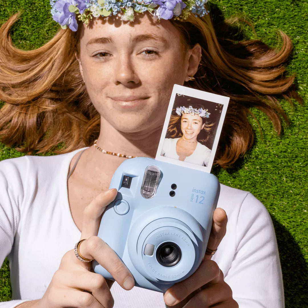 Sfeerafbeelding instax mini 12™ camera Pastel Blue waarin een dame, die ligt op het gras, de camera vasthoud en er een selfie uitkomt. 