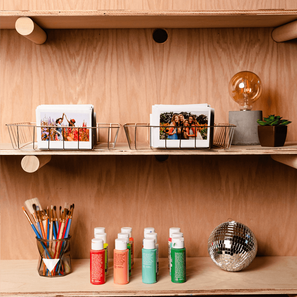 Sfeerafbeelding instax™ WIDE film, waarin de foto's in een mandje op een plank staan. Verder zijn er knutselspulletjes zichtbaar, de foto geeft een knusse sfeer.