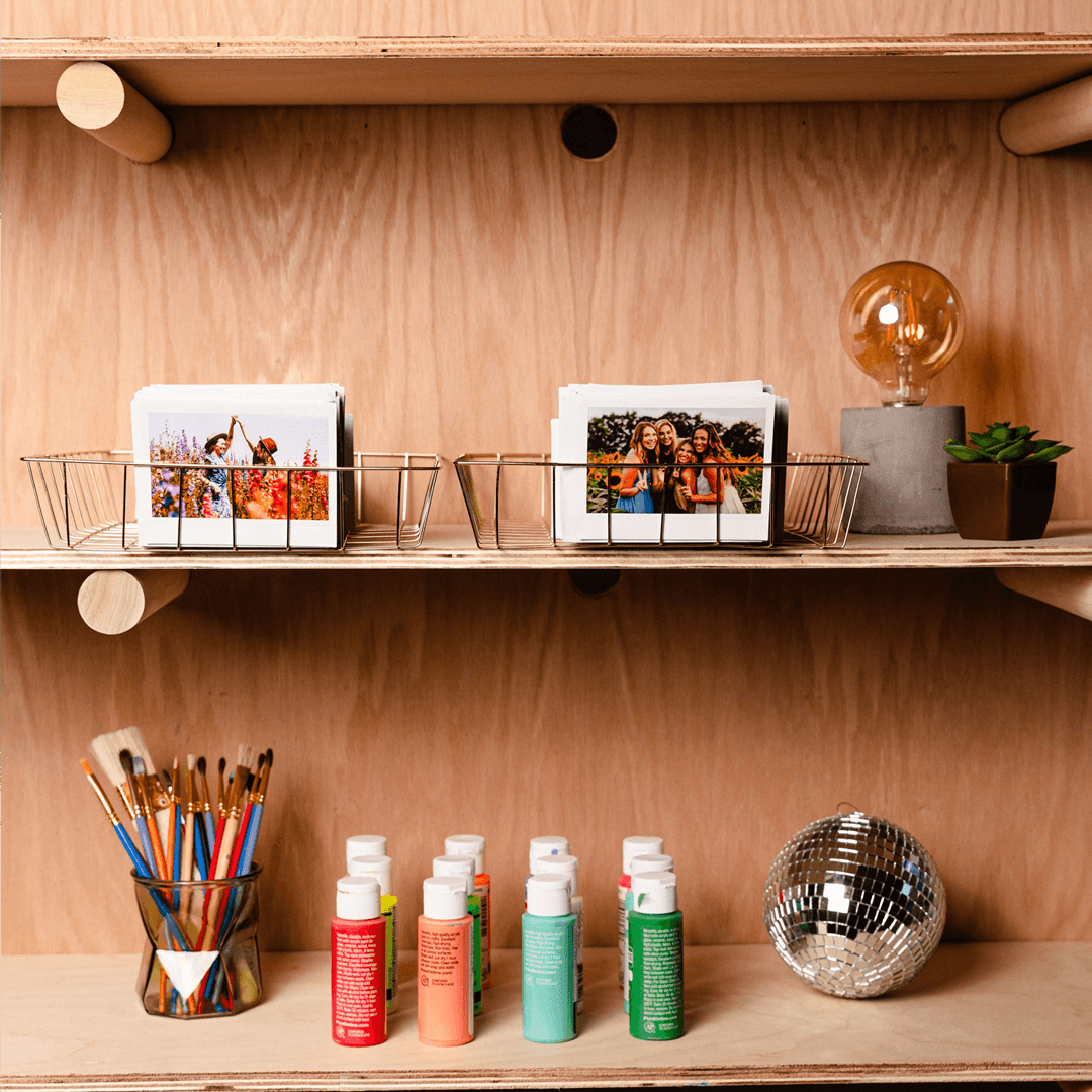 Sfeerafbeelding instax™ WIDE film, waarin de foto's in een mandje op een plank staan. Verder zijn er knutselspulletjes zichtbaar, de foto geeft een knusse sfeer.