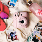 Sfeerafbeelding instax mini 12™ camera Blossom Pink. Op de foto ligt de camera op een doek, met daarom heen zomerse accessoires: slippers, zonnebril, waterfles en verschillende instax™ foto's