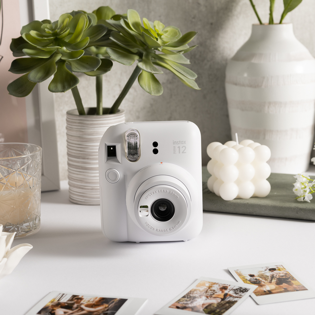 Sfeerafbeelding instax mini 12™ camera Clay White.  De camera staat op een tafel met meerdere accessoires: een plant, een glas met sap, een plank met een kaars en losse foto's.