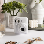 Sfeerafbeelding instax mini 12™ camera Clay White.  De camera staat op een tafel met meerdere accessoires: een plant, een glas met sap, een plank met een kaars en losse foto's.