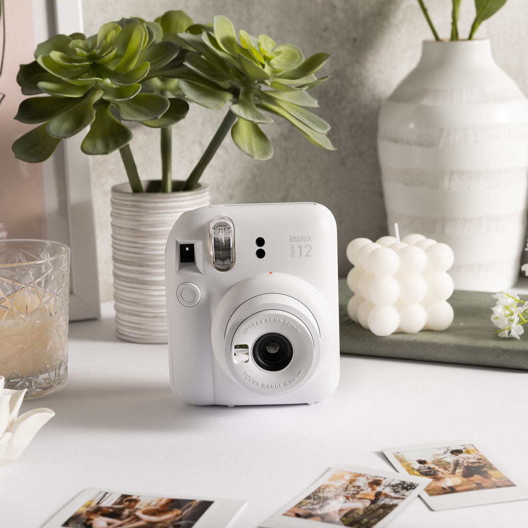 Sfeerafbeelding instax mini 12™ camera Clay White.  De camera staat op een tafel met meerdere accessoires: een plant, een glas met sap, een plank met een kaars en losse foto's.
