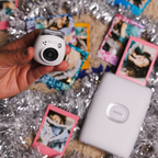 instax Pal™ camera Milky White met instax mini Link 2 printer in Wit. Uit de printer komen foto's en rondom de printer en de camera liggen feestelijke accessoires: foto's en slingers. 