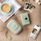 Instax mini LiPlay matcha green vooraanzicht, op een bank met daaromheen de instax mini LiPlay matcha green case, een kop koffie, de Instax LiPlay app en een aantal foto's