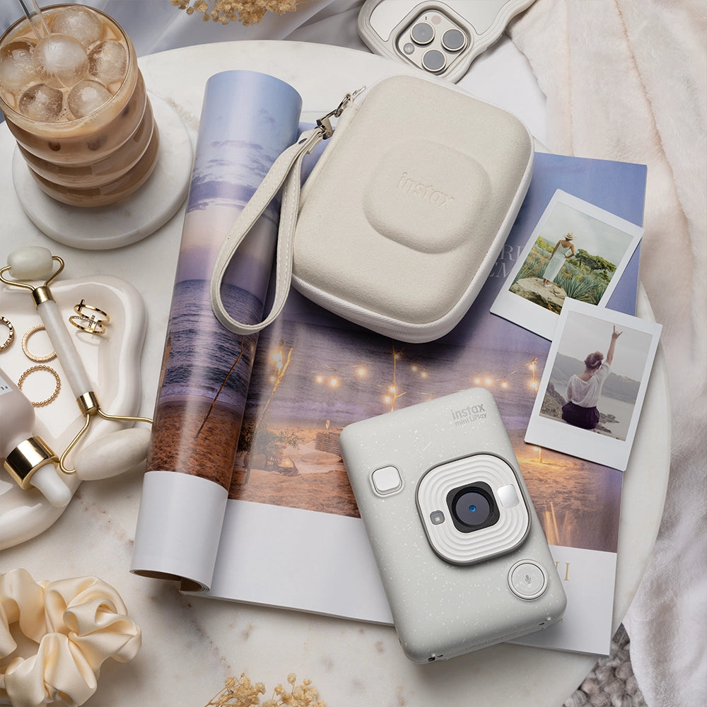 Instax mini LiPlay Misty white op een bijzettafel met daaronder een magazine, om de camera liggen foto's een instax mini LiPlay misty white case, sieraden en ijskoffie