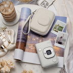 Instax mini LiPlay Misty white op een bijzettafel met daaronder een magazine, om de camera liggen foto's een instax mini LiPlay misty white case, sieraden en ijskoffie